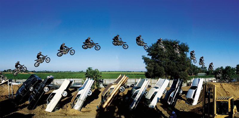 A freestyle motocross rider shown in multiple exposures arcing over a row of Cadillacs buried nose-down in the dirt
