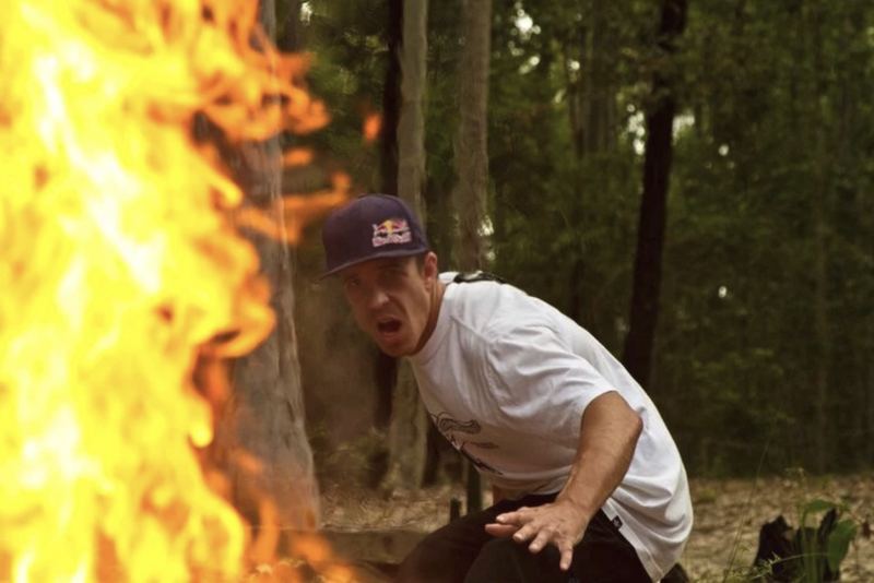 A man in a Red Bull cap crouches beside a large fire in a forest