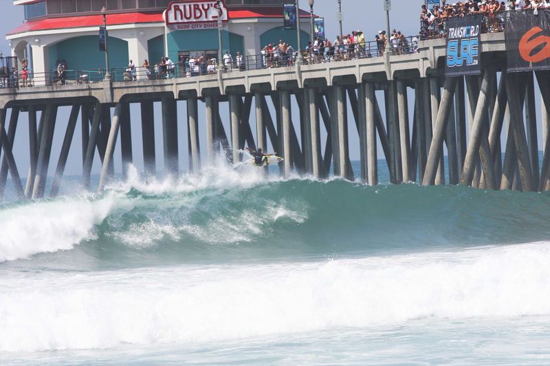 A surfer launches off a wave beside the Huntington Beach pier during the US Open of Surfing, with spectators lining the pier