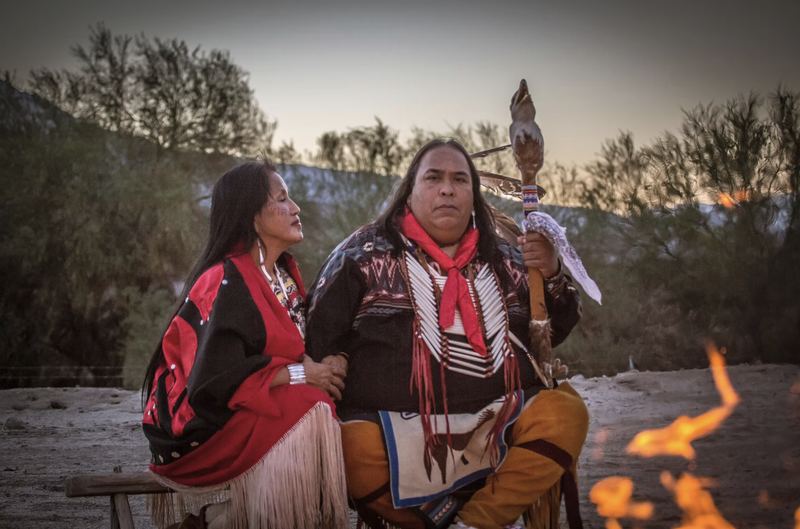 Two people in traditional regalia seated by a fire at dusk in the desert