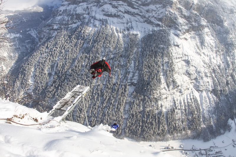 A wingsuit flyer launches from a snowy cliff platform above an alpine valley in Switzerland